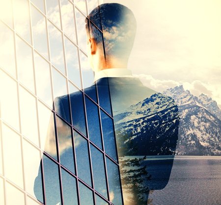Geschäftsmann steht mit dem Rücken zum Betrachter vor Glasfassade und Gebirge, Doppelbelichtung